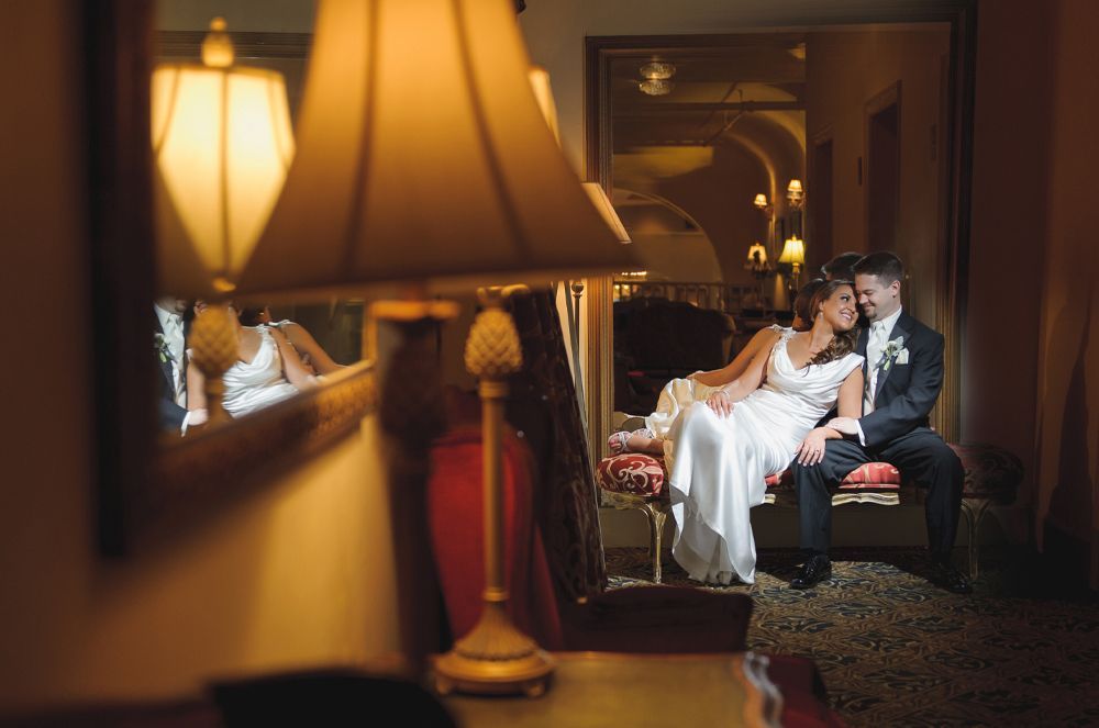 Bride and groom sit on a bench, smiling, in a warmly lit hallway. A lamp is in the foreground.