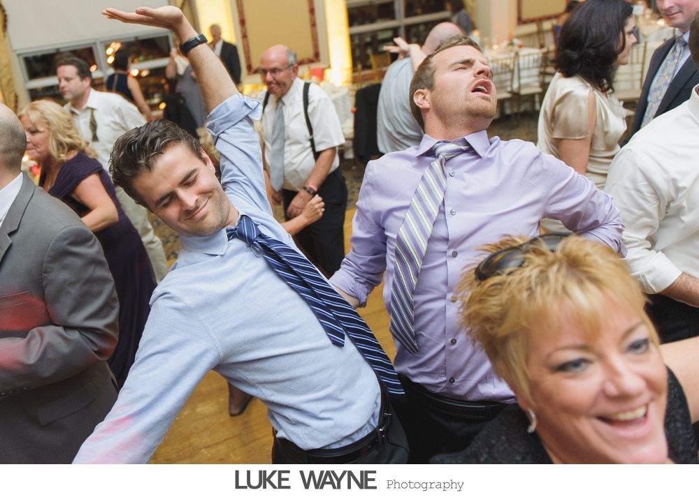 Two men enthusiastically dancing, arms outstretched, at a wedding reception. Other guests nearby, festive atmosphere.