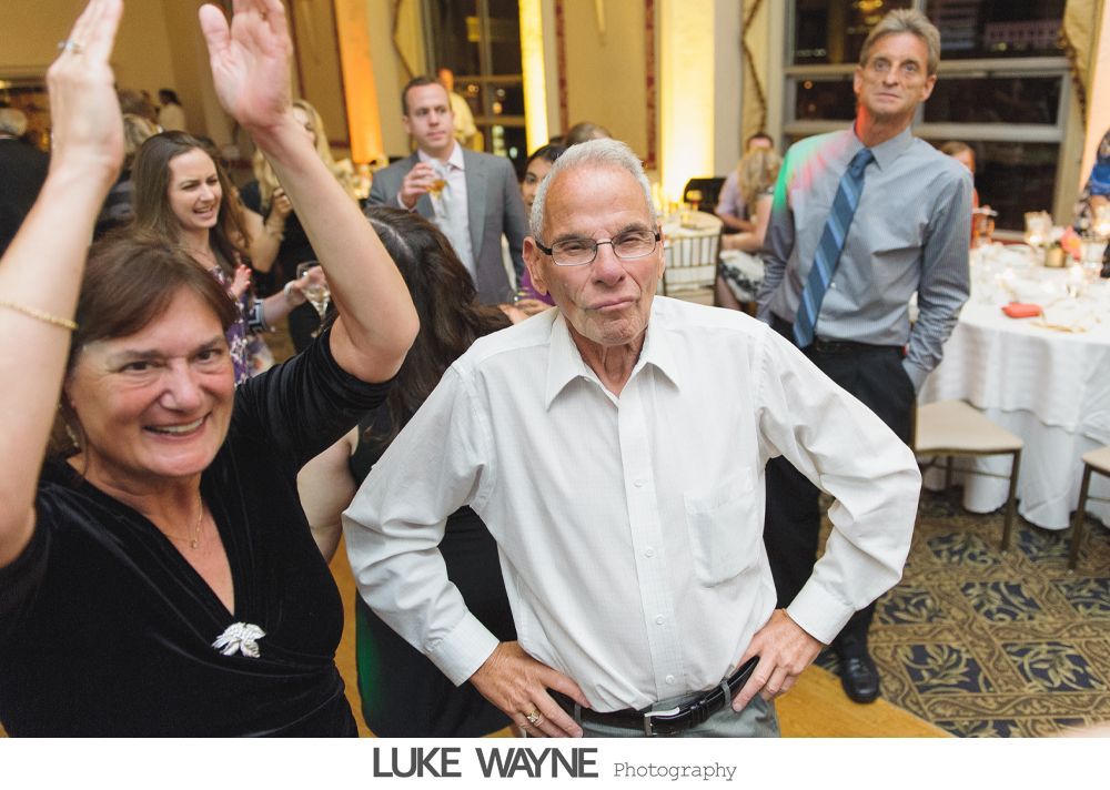 People dancing at a party; woman raising hands, man with hands on hips, another man watching, indoor setting.