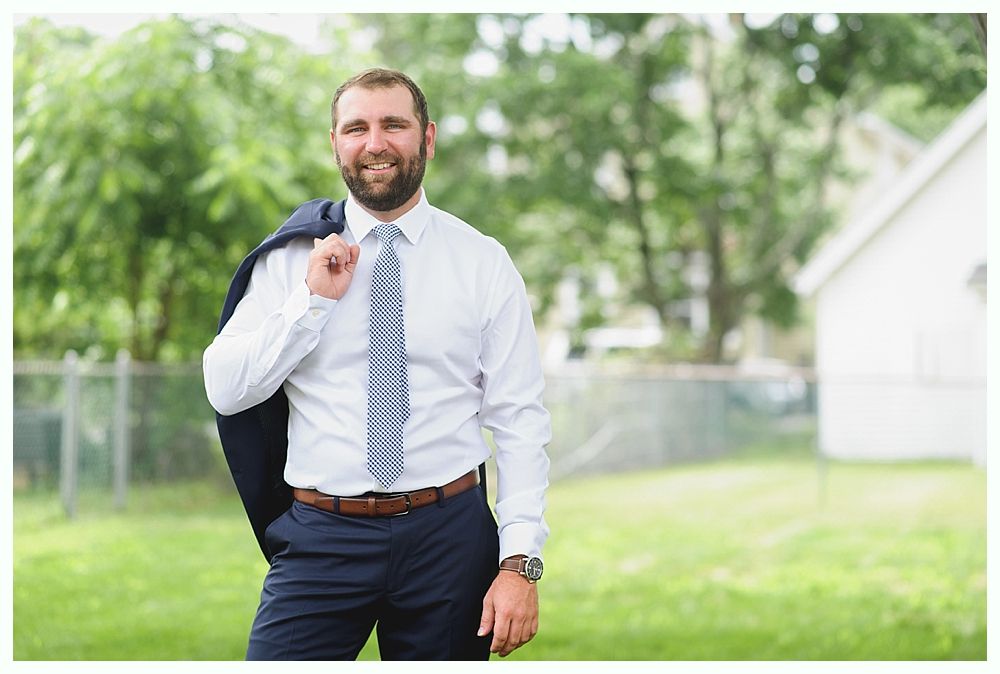 Man in white shirt, tie, and navy suit, smiling, holding coat over shoulder in a grassy yard.