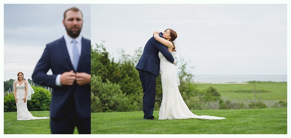 Wedding couple embraces on a grassy hill; man in blue suit, woman in white dress, ocean in background.