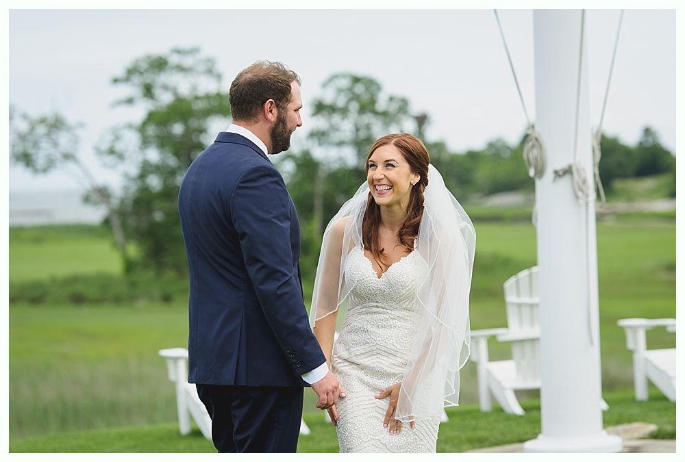 Bride and groom smiling at each other during an outdoor wedding ceremony. Bride wears a white gown and veil. Groom in a blue suit.