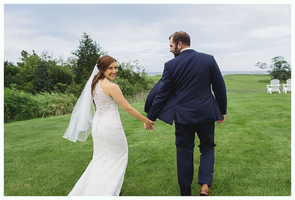 Bride and groom holding hands, walking on grass, bride looking at camera, overlooking water.