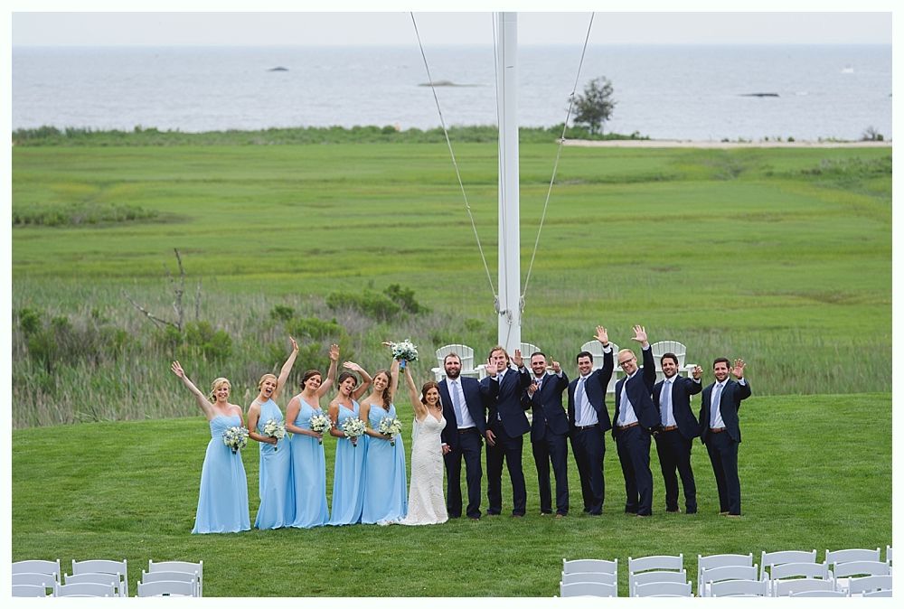 Wedding party poses outdoors with ocean view; women in blue, men in suits, arms raised.