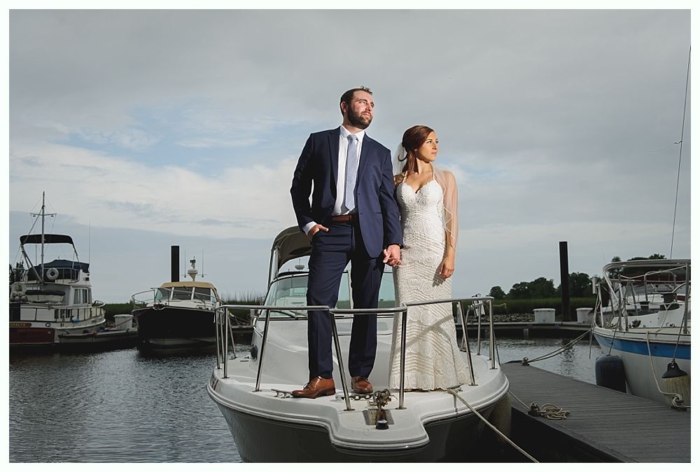 Couple in wedding attire on a boat in a harbor, looking off into the distance. Boats and a dock are in the background.