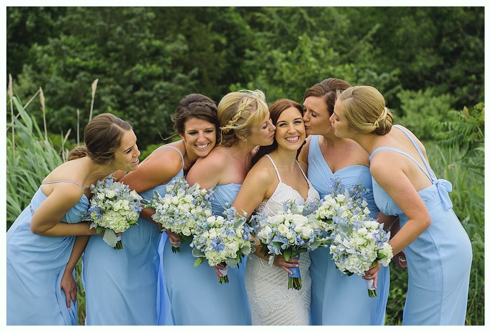 Bride with bridesmaids in light blue dresses, all smiling with bouquets; outdoors.