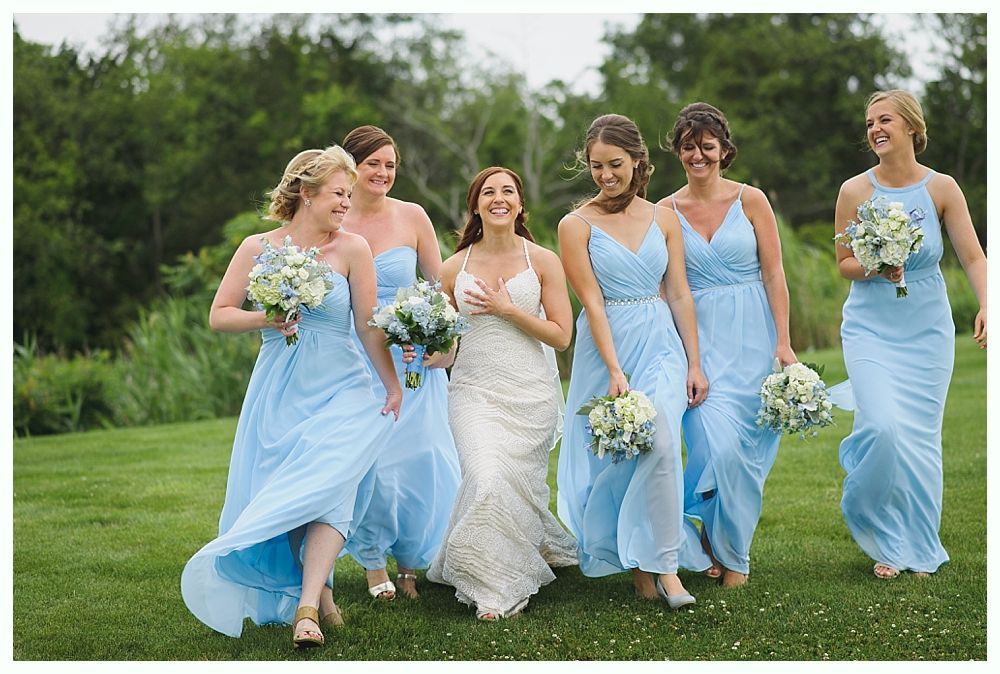 Bride in white gown walks with bridesmaids in light blue dresses, smiling on a grassy field.