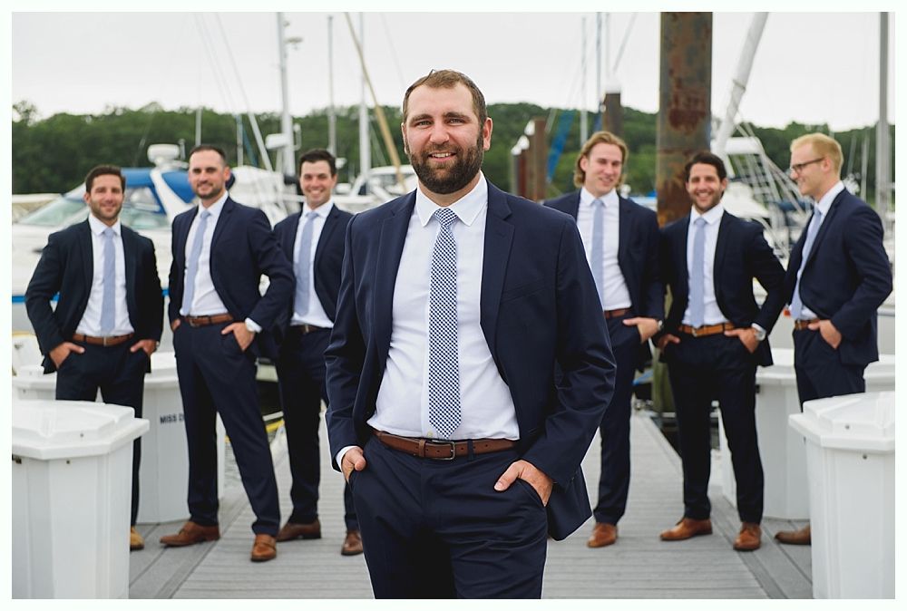 Men in suits on a dock, one smiling at camera, others in the background. Boats in the marina.