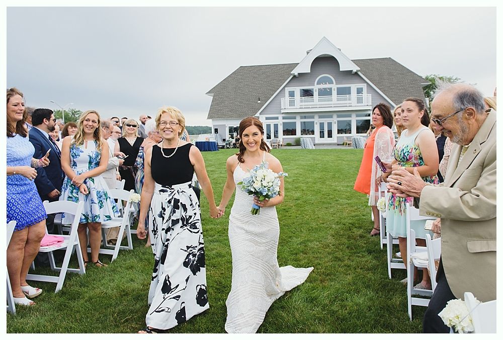 Bride and woman walk down an aisle, a house in the background. Guests clap.