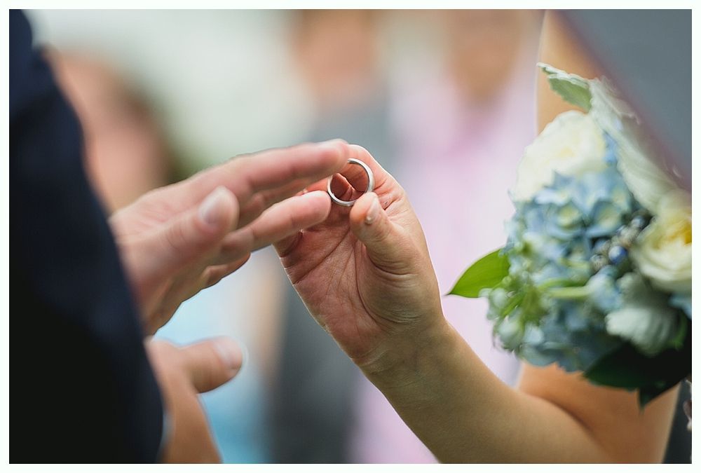 Wedding ring exchange; hands holding a silver ring near a bouquet of white and blue flowers.