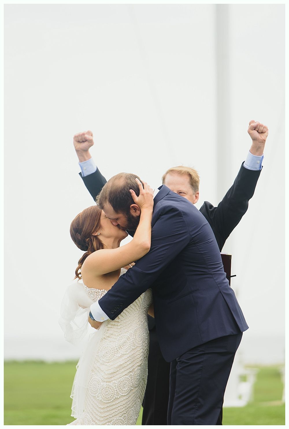 Bride and groom kissing during ceremony, officiant cheering with arms raised; outdoors, white background.