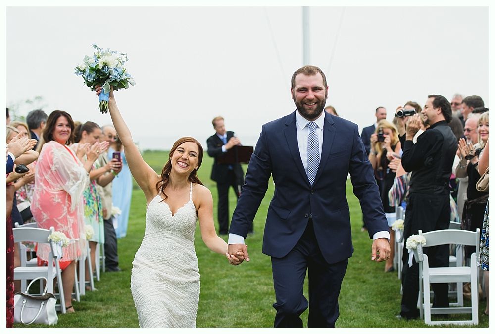 Newlyweds walking, holding hands, and smiling after the ceremony, outdoors with guests clapping.
