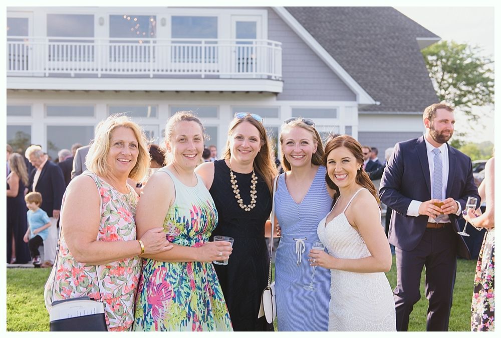 Five women smiling together outdoors at a wedding reception; a two-story building in the background.