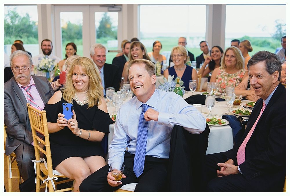 Wedding reception: Guests laughing at a table. Woman holds phone, man raises a glass, set in a bright room.