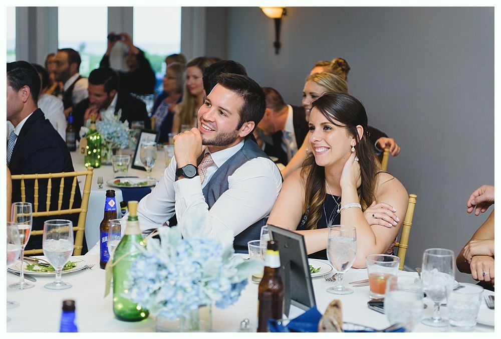 Couple smiling at a wedding reception; tables with centerpieces, guests in background.