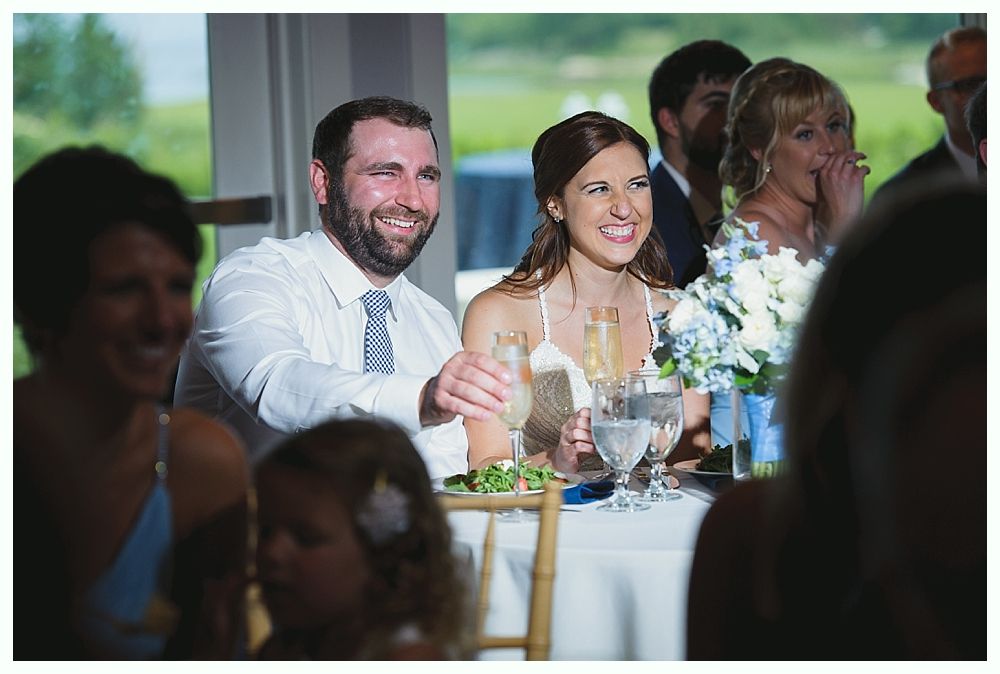 Bride and groom smiling at a wedding reception. They're seated at a table with guests and flowers.