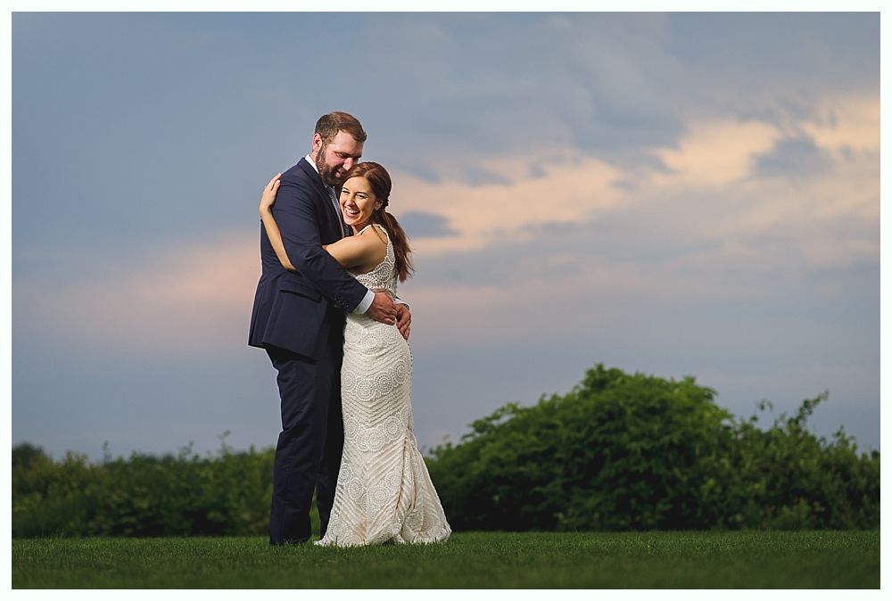 Couple embracing outdoors, woman in white wedding dress, man in blue suit, smiling. Sky and greenery in background.