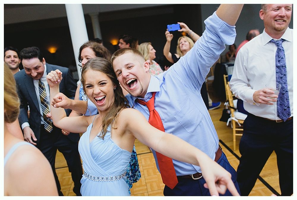 People dancing and celebrating at an event. A woman in a blue dress and a man in a blue shirt with a red tie are smiling and looking at the camera.
