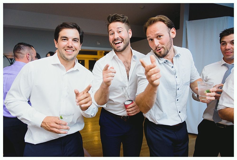 Three men smile and point towards the camera at an event, one holding a drink.