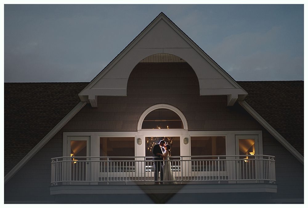 Couple on a balcony embracing, lit from within, under the eaves of a building at dusk.