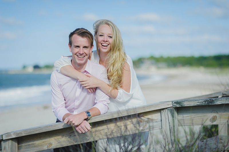 Couple holding hands in a field, woman twirling. They wear formal clothing, trees in the background, sunny day.