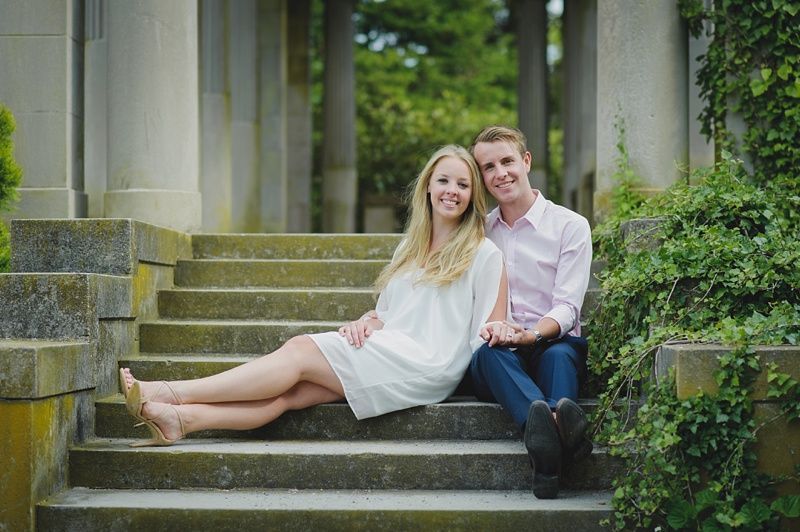 Couple in formal attire posing outdoors. Woman in red and black dress, man in black suit, holding hands, smiling.