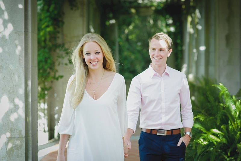 Couple walking and smiling; woman in white dress, man in pink shirt; outdoor setting with columns and greenery.