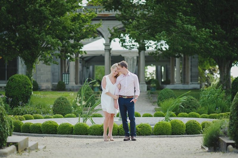 Couple embraces on a grassy lawn with a house in the background at sunset.