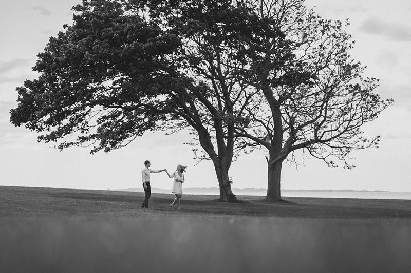 Couple embraces on a grassy hill in front of a large house, sun shining.