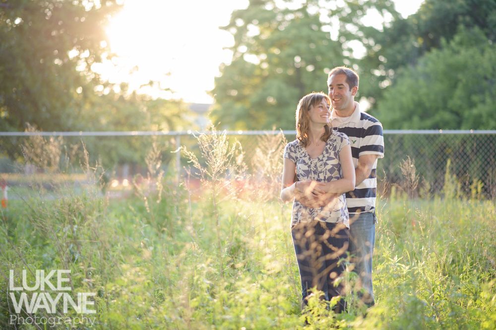 Couple embracing in a field of tall grass, sunlight streaming through. Man in striped shirt. Woman in floral dress.