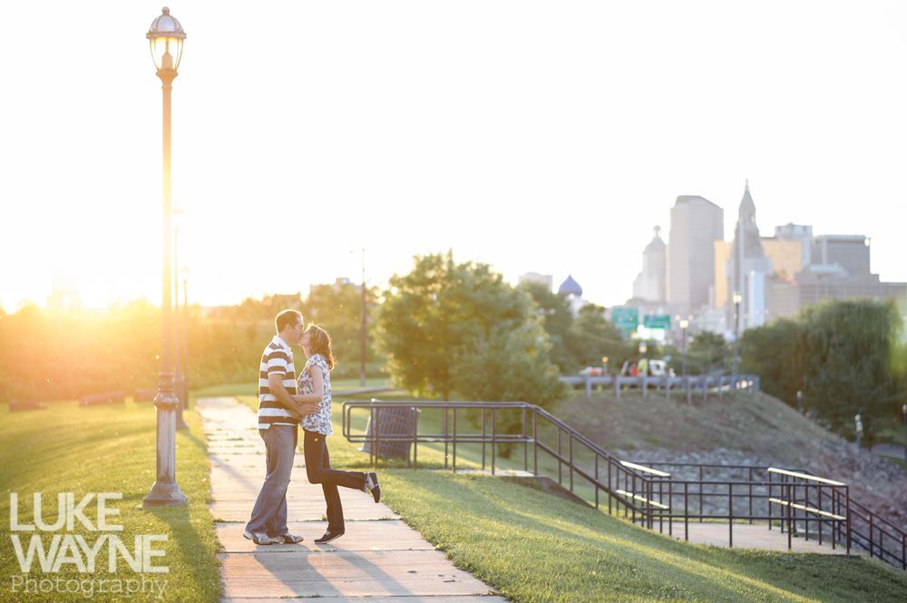 Couple kissing on a path at sunset, city skyline in background.