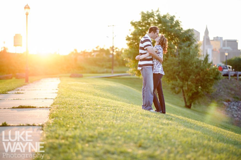 Couple kissing on grassy hill at sunset, city skyline in background.