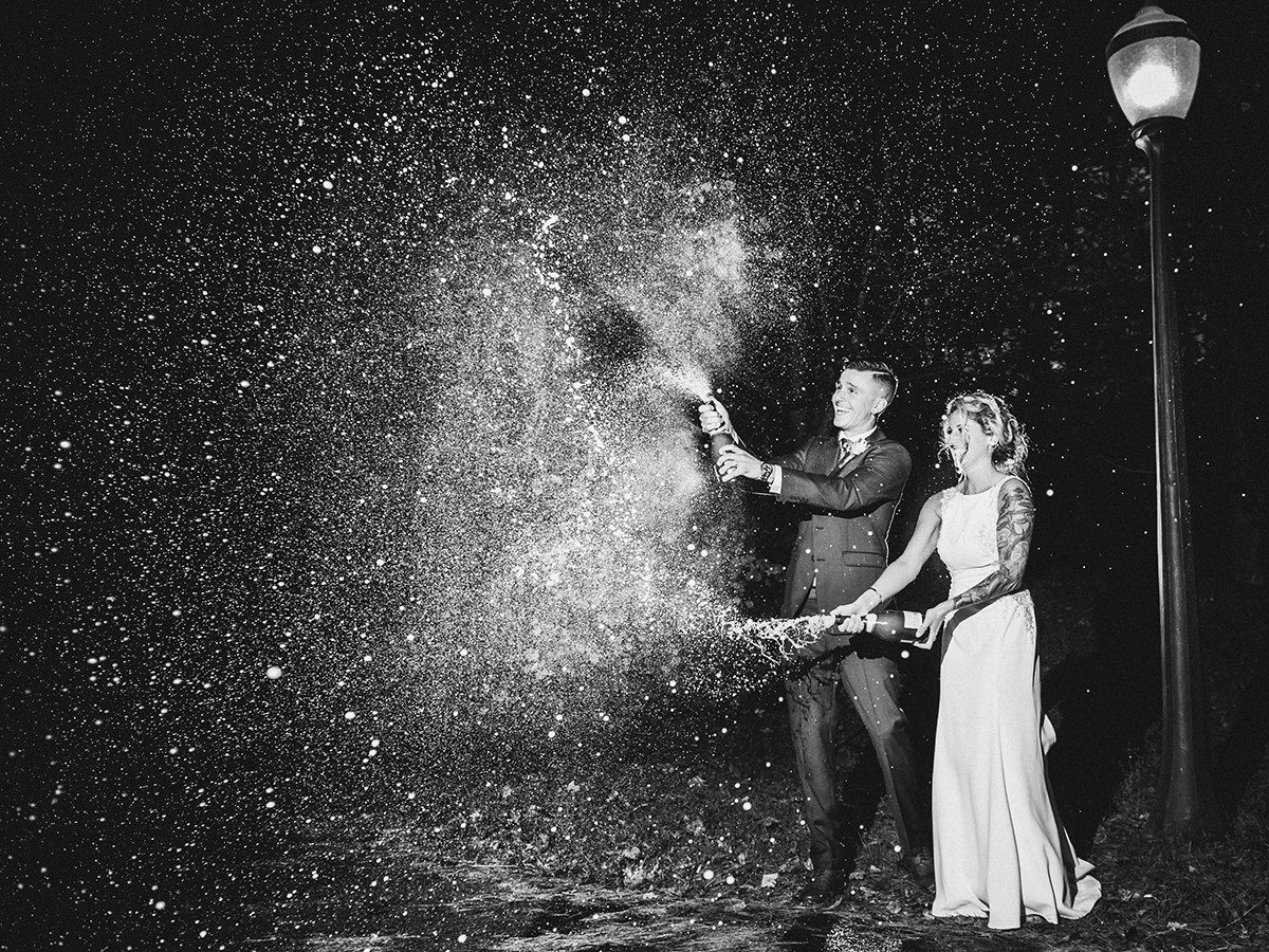 Newlyweds pop champagne, spraying bubbles in the night. A lamppost illuminates the scene.