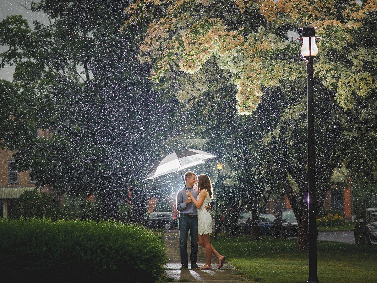 Couple under umbrella in the rain, embracing under a street lamp in front of a tree.