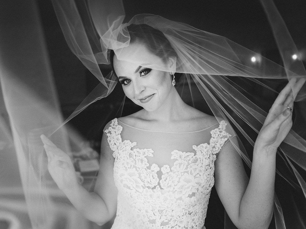 Bride in lace wedding dress, veil billowing, looking at camera and smiling.