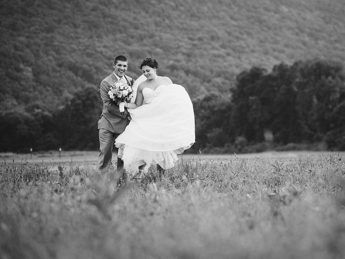 Bride and groom laugh, running through a field with a mountain backdrop. Black and white.