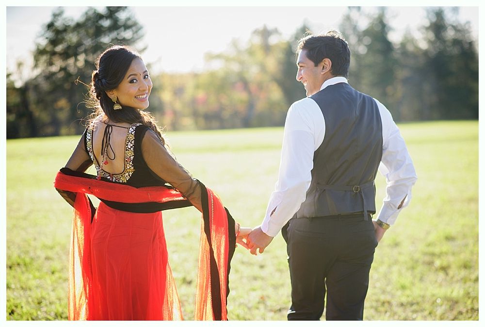 Couple holding hands in a field, woman in red and black outfit looking back, man in a vest.