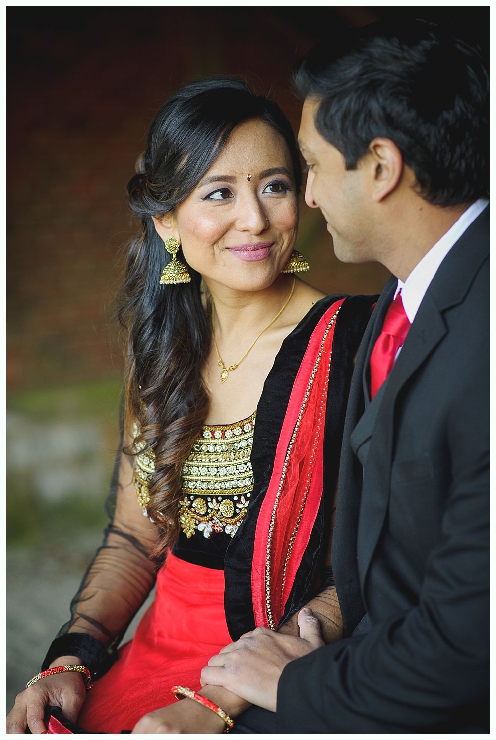 Couple in formal wear looking at each other; woman wearing red sari and gold earrings, man in a black suit.