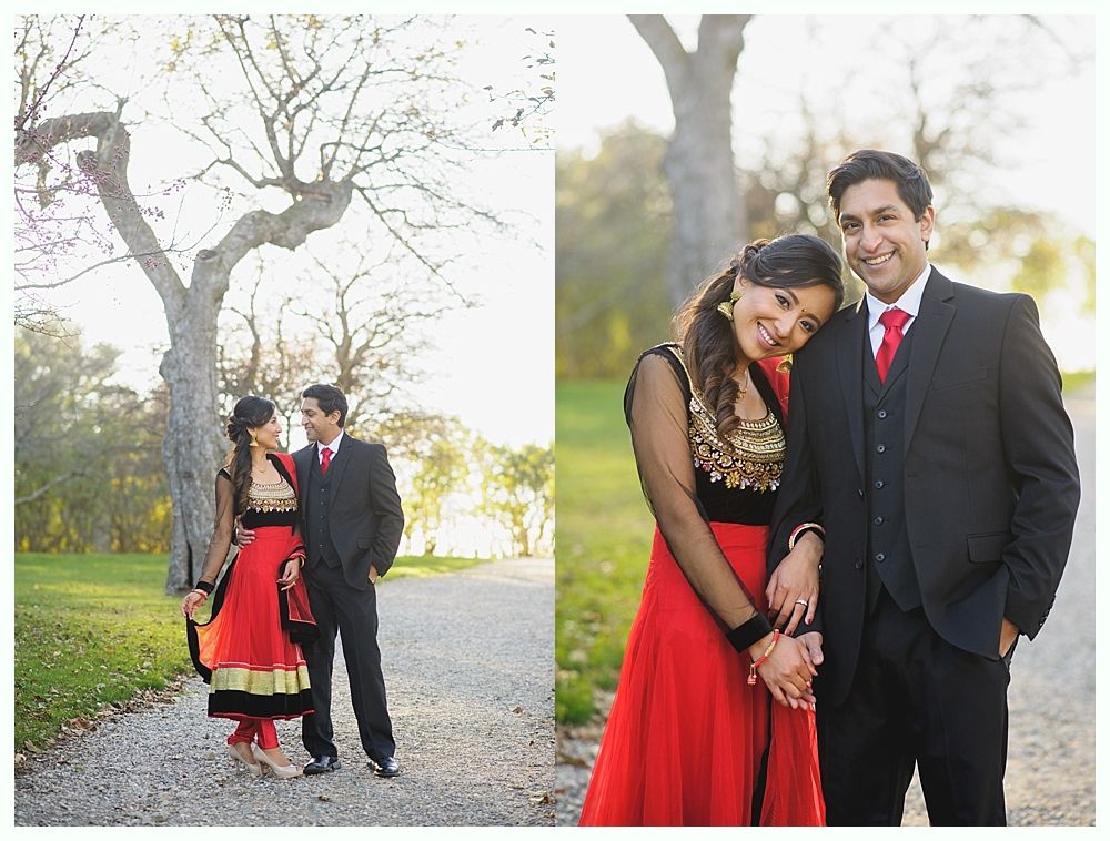 Couple in formal attire posing outdoors. Woman in red and black dress, man in black suit, holding hands, smiling.