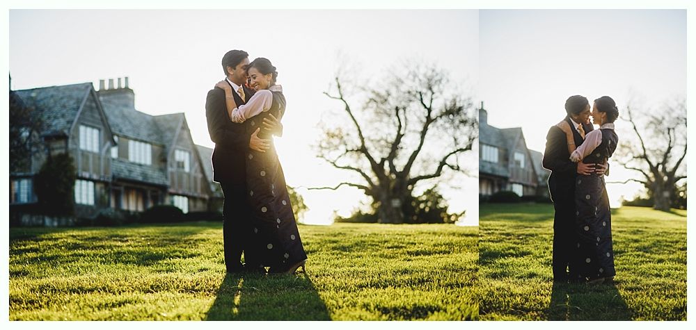 Couple embraces on a grassy hill in front of a house, backlit by the sun.