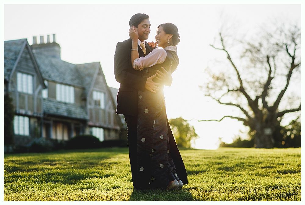 Couple embraces on a grassy lawn with a house in the background at sunset.