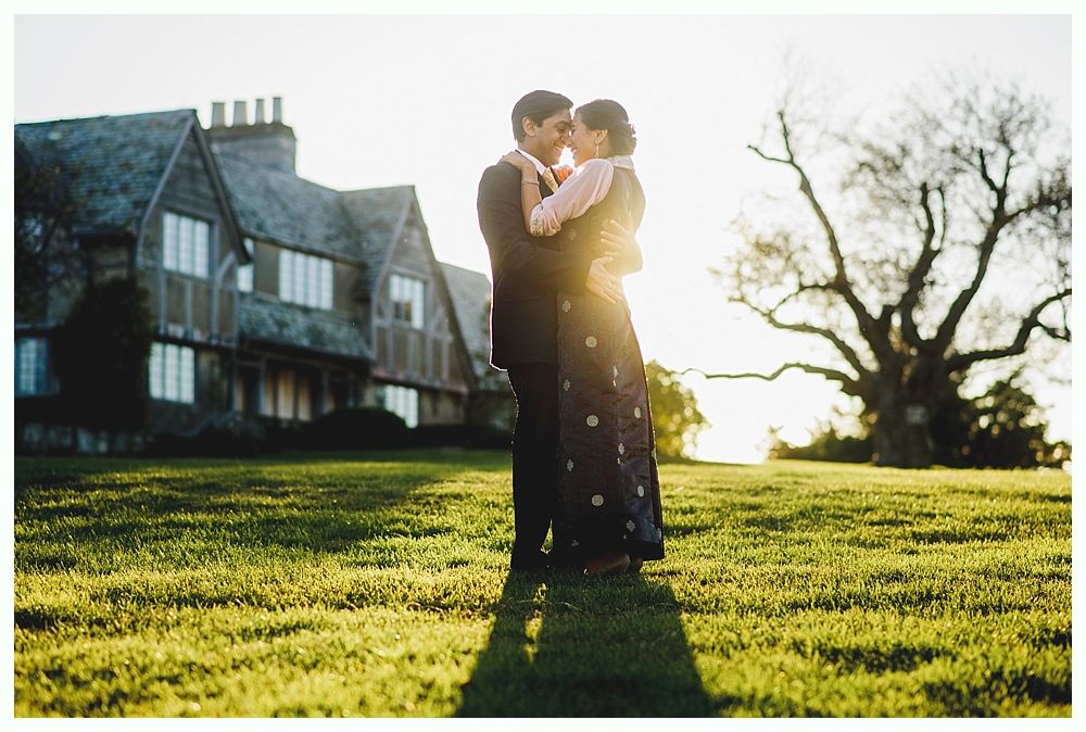 Couple embraces on a grassy hill in front of a large house, sun shining.
