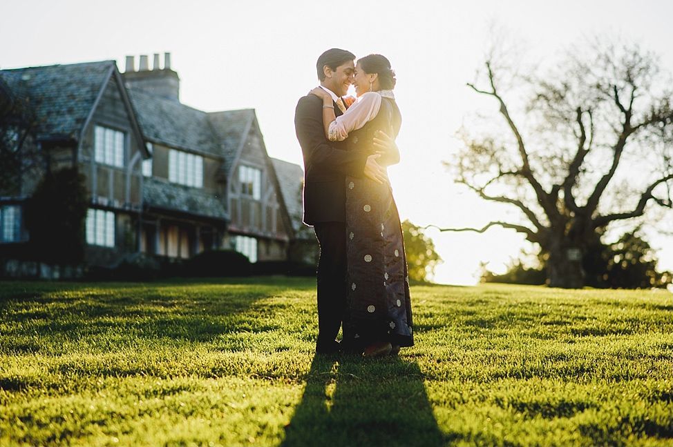 Couple embracing outdoors with a large house in the background at sunset.