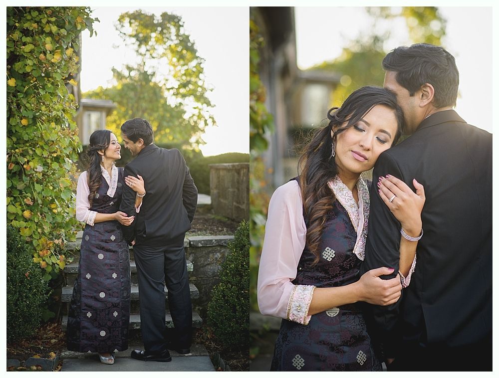 Couple in traditional attire embraces outdoors, smiling. Dark jackets, stone steps, foliage.