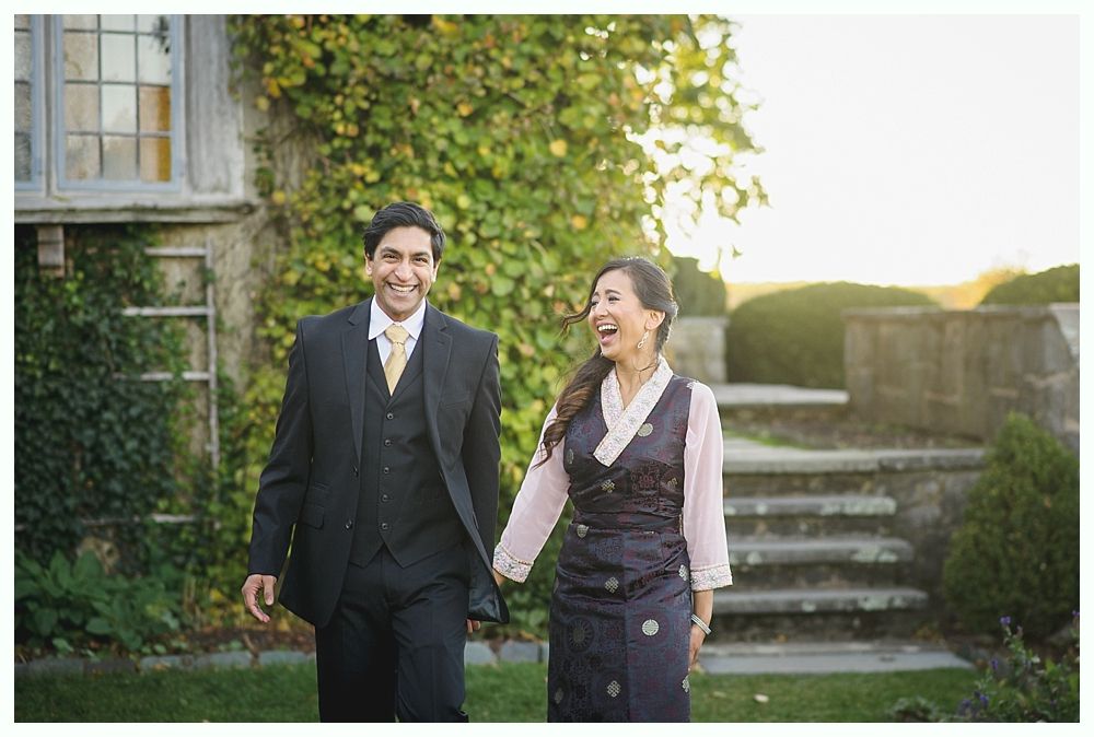 Couple holding hands, smiling while walking outside. Man wears suit, woman wears traditional dress. Ivy-covered building in background.