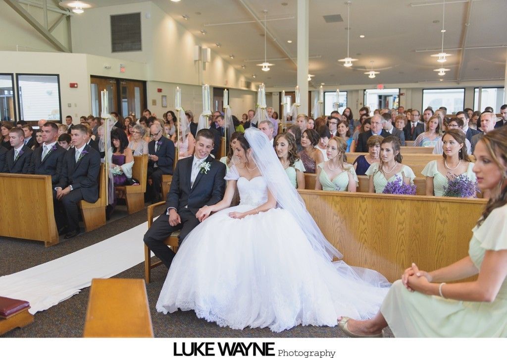 Bride and groom seated in church, ceremony in progress. Guests in pews. White carpet, long train.