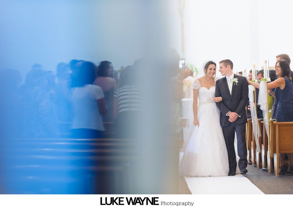 Bride and groom walking down aisle. Bride in white dress, groom in suit. Guests watch.