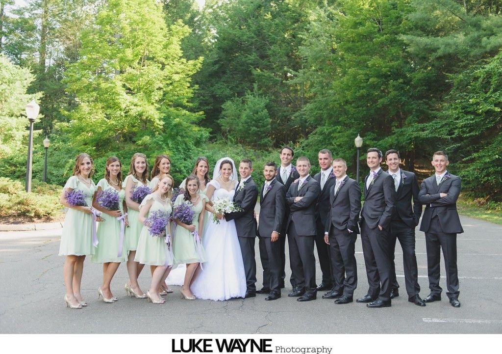 Wedding party poses on pavement, bride in white dress, bridesmaids in green dresses, groomsmen in black suits, trees in background.