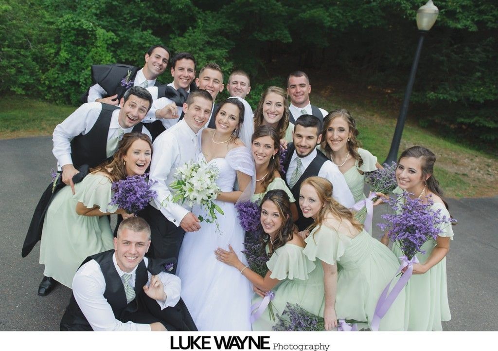 Wedding party poses outdoors. Bride and groom surrounded by bridesmaids in green and groomsmen in black vests.