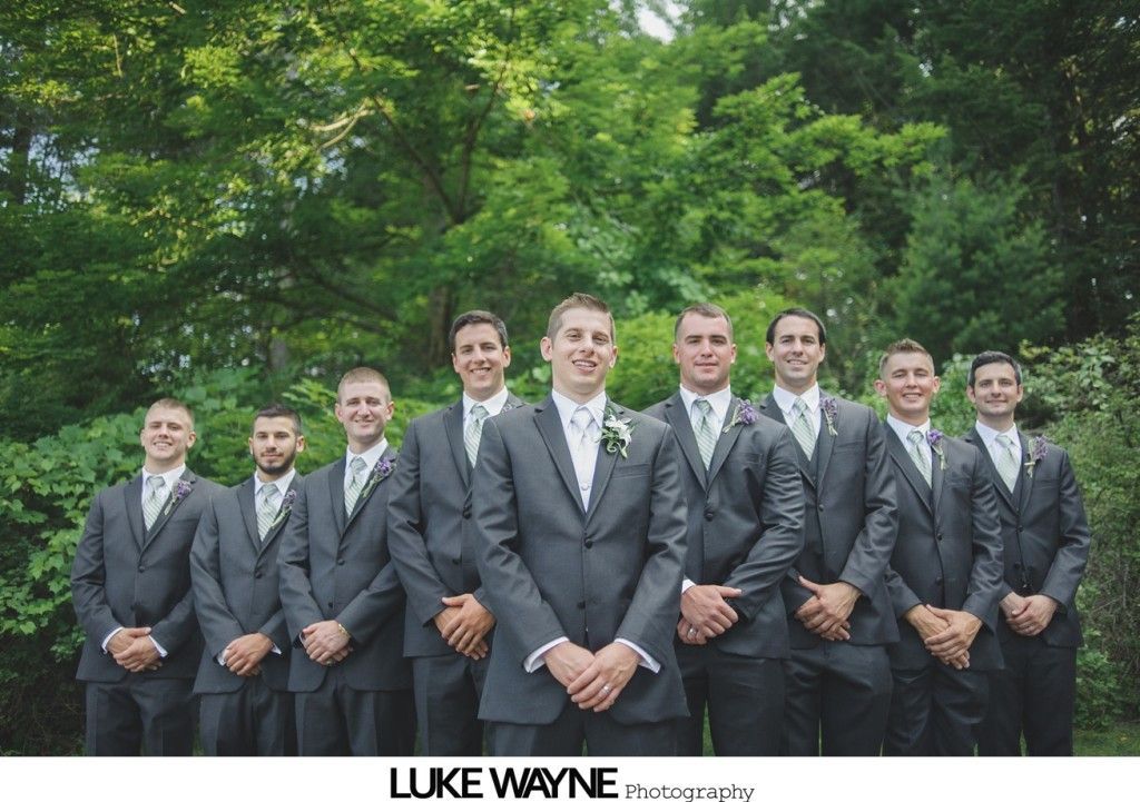 Groomsmen in gray suits pose outdoors in front of lush green trees, arms crossed, smiling slightly.